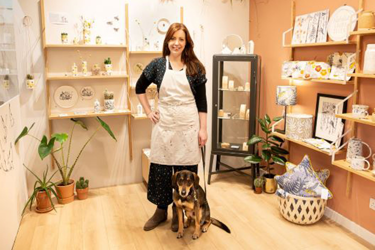 A woman in an apron standing in her studio with her dog, surrounded by ceramics and homewares
