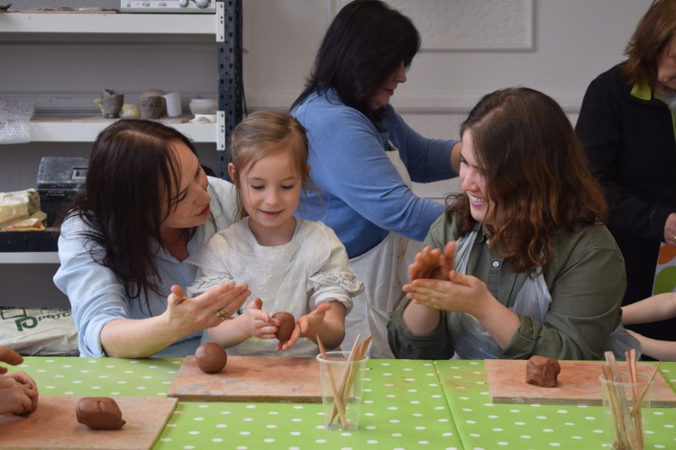 A child and two adults work on some clay at a table covered in craft materials