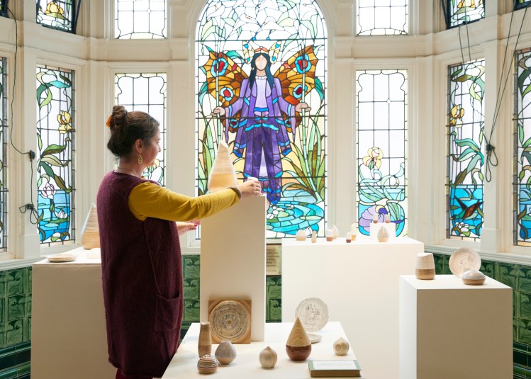 A tiled room with Arts and Crafts era stained glass window contains a display of craft and a maker setting up her work